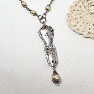 May include: A silver necklace featuring a vintage spoon charm engraved with the letter "C" and a pearl drop. The necklace is crafted with a silver chain and white pearls.