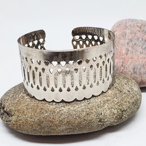 May include: Silver metal bracelet with a decorative cut-out design. The bracelet is resting on two gray rocks.