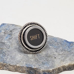 May include: A silver ring with a black and silver round top. The top has a black background with the word "SHIFT" in gold lettering.