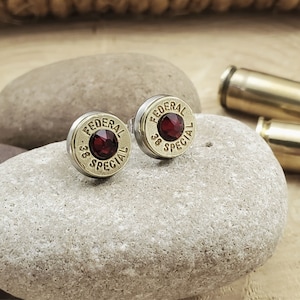 May include: A pair of gold-toned earrings featuring a red gemstone set in a bullet casing with the text "FEDERAL 38 SPECIAL".
