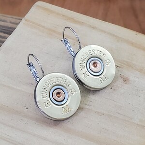 May include: A pair of gold-toned earrings featuring a 20 gauge Winchester AA shotgun shell. The earrings have a silver-toned hook closure.
