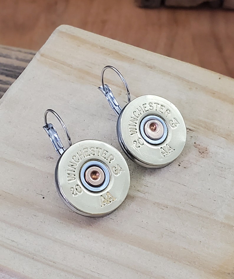 May include: A pair of gold-toned earrings featuring a 20 gauge Winchester AA shotgun shell. The earrings have a silver-toned hook closure.