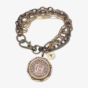Könnte beinhalten: Ein mehrsträngiges Armband mit verschiedenen Ketten in Gold-, Bronze- und Silbertönen. Ein großer, runder Anhänger mit dem Text "CANTON CITY LINES INC" und dem Buchstaben "C" hängt am Armband. Ein kleiner Anhänger mit dem Buchstaben "A" ist ebenfalls angebracht.