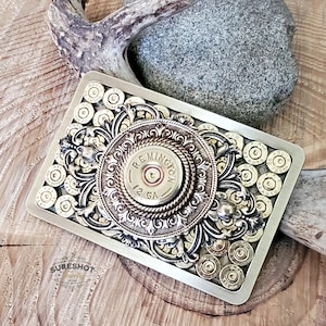 May include: A rectangular belt buckle featuring a central Remington 12 GA shotgun shell casing surrounded by ornate silver-toned filigree metalwork. The background is filled with an array of various brass-colored bullet casings.