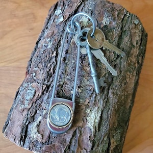 May include: A large silver-toned metal safety pin keychain featuring a circular coin charm with a buffalo design and a twisted gold-toned border. The pin holds two silver-toned metal keys and one vintage-style skeleton key on a metal ring.