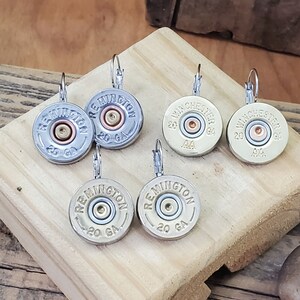 May include: Six pairs of silver and gold tone earrings featuring shotgun shell casings with the words "Remington" and "Winchester" printed on them. The casings are 20 gauge.