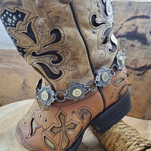 May include: A pair of brown leather cowboy boots with intricate cutouts and silver accents. The boots have a silver chain with bullet casings attached to the top of the boot.