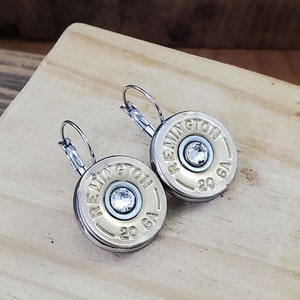 May include: A pair of silver earrings with a gold-coloured round charm that says "REMINNGTON 20 GA" with a clear rhinestone in the centre.