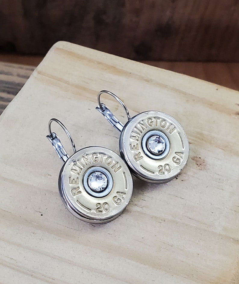 May include: A pair of silver earrings with a gold-coloured round charm that says "REMINNGTON 20 GA" with a clear rhinestone in the centre.
