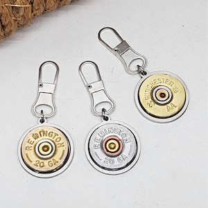 May include: Three metal zipper pulls with circular charms. The charms feature the text "Remington 20 GA" and "Winchester AA 20 GA" around a central brass-colored circle. The zipper pulls are silver-toned.