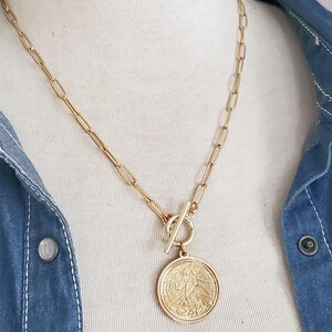 May include: A gold chain necklace with a large, round pendant featuring an angel design. The pendant is attached to the chain with a toggle clasp.