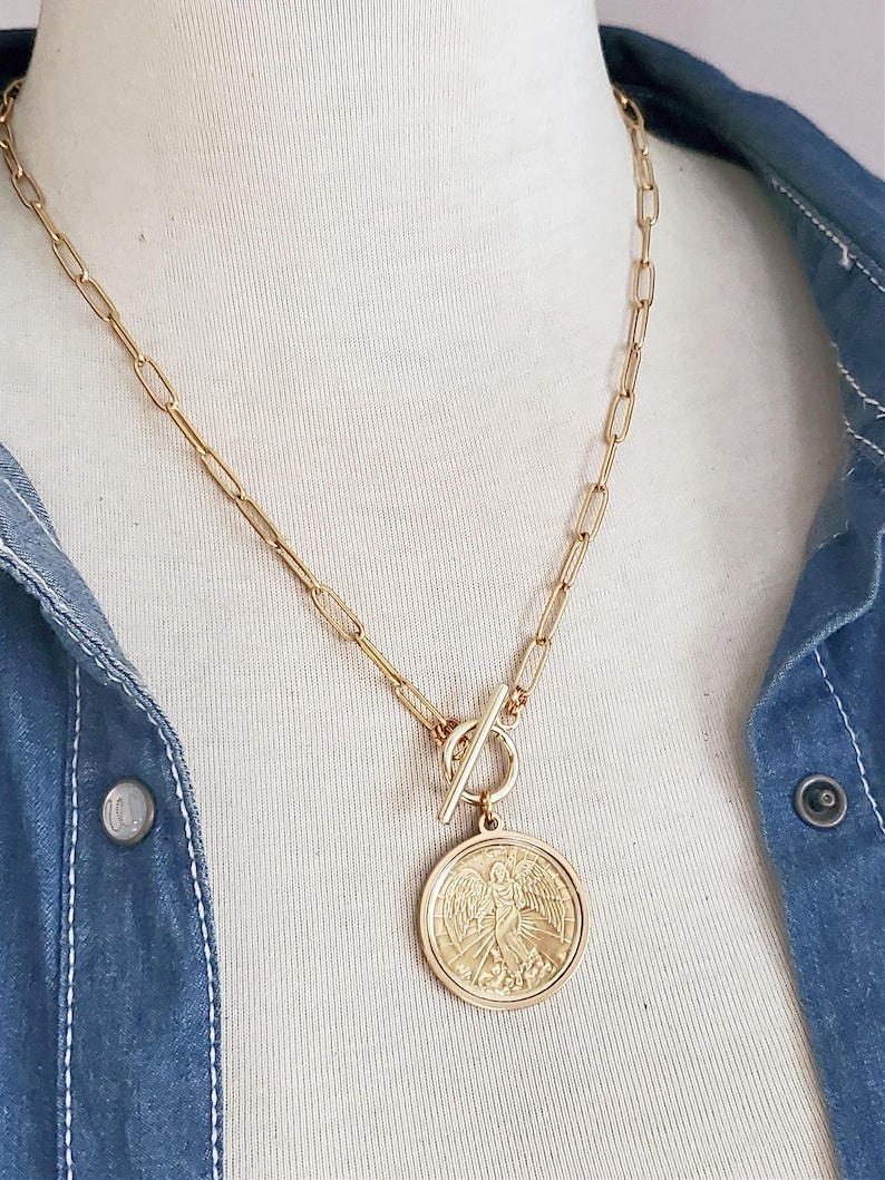 May include: A gold chain necklace with a large, round pendant featuring an angel design. The pendant is attached to the chain with a toggle clasp.