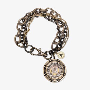 Puede incluir: Pulsera de múltiples cadenas en tonos bronce y negro. Incluye un colgante circular grande con el texto "BEAVER VALLEY MOTOR COACH CO." y un pequeño dije. Con cierre de mosquetón.