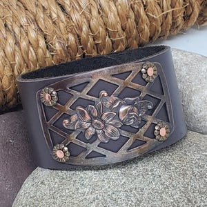 Puede incluir: Brazalete de cuero marrón oscuro con una placa metálica decorativa. La placa presenta un diseño floral con una rosa y un girasol, dentro de una rejilla de diamantes. Pequeños detalles de color cobre en los bordes.