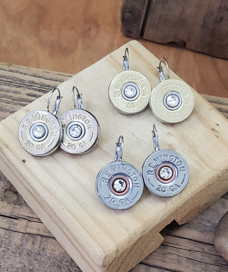 May include: A pair of silver earrings with a clear rhinestone in the centre of each earring. The earrings are shaped like shotgun shells with the words "Remington" and "20 GA" printed on them. There is also a pair of gold earrings with the words "Winchester" and "20 GA" printed on them.