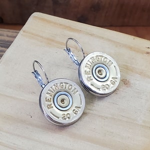 May include: A pair of silver hoop earrings with a gold-coloured 20 gauge shotgun shell charm. The charms have the text "Remington" printed on them.