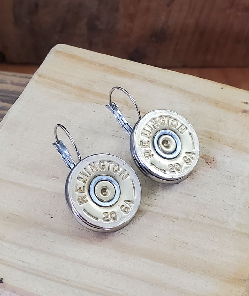May include: A pair of silver hoop earrings with a gold-coloured 20 gauge shotgun shell charm. The charms have the text "Remington" printed on them.