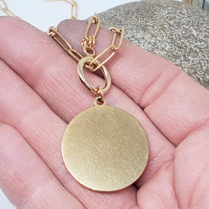 May include: The back of a gold-toned pendant with a circular shape. The pendant is attached to a gold chain.