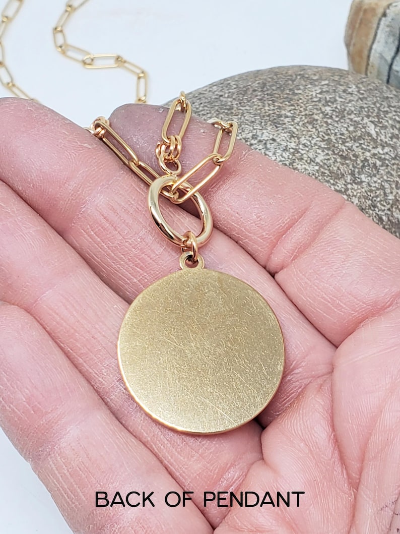 May include: The back of a gold-toned pendant with a circular shape. The pendant is attached to a gold chain.