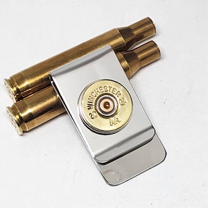 Może przedstawiać: Srebrny klips na pieniądze ze złotą łuską po naboju Winchester 20 GA AA. Dwie dodatkowe mosiężne łuski po nabojach znajdują się za klipsem. Stylowy dodatek.