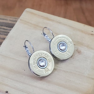 May include: A pair of silver hoop earrings with a gold-coloured bullet casing charm. Each charm has a clear rhinestone in the centre and the text "WINCHESTER 20 GA AA" printed on the side.