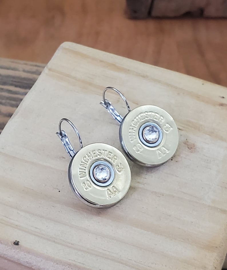 May include: A pair of silver hoop earrings with a gold-coloured bullet casing charm. Each charm has a clear rhinestone in the centre and the text "WINCHESTER 20 GA AA" printed on the side.