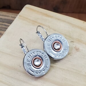 May include: A pair of silver earrings with a clear rhinestone in the centre of each earring. The earrings are made from repurposed shotgun shells with the text "REMMINGTON 20 GA" printed on each shell.