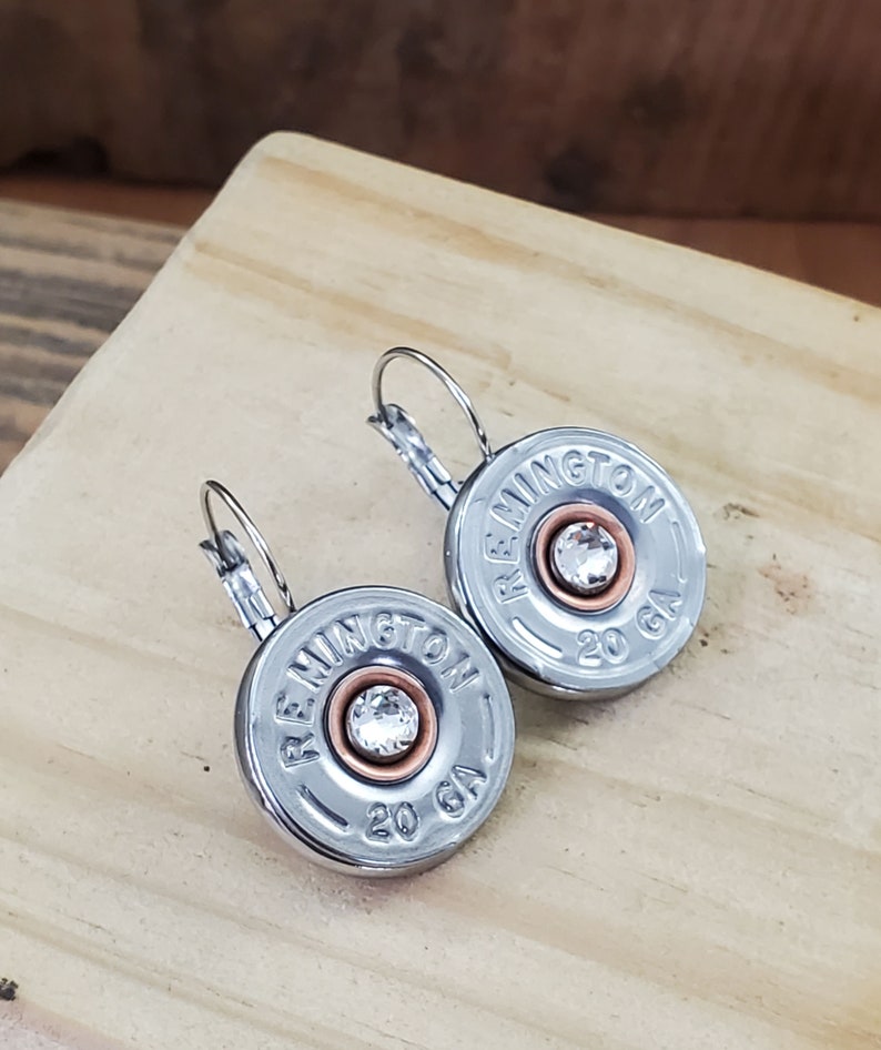May include: A pair of silver earrings with a clear rhinestone in the centre of each earring. The earrings are made from repurposed shotgun shells with the text "REMMINGTON 20 GA" printed on each shell.