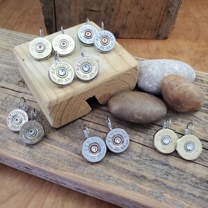 May include: A set of silver and gold tone earrings featuring a round, silver-coloured shotgun shell with a clear rhinestone in the centre. The earrings are displayed on a wooden surface with small rocks.