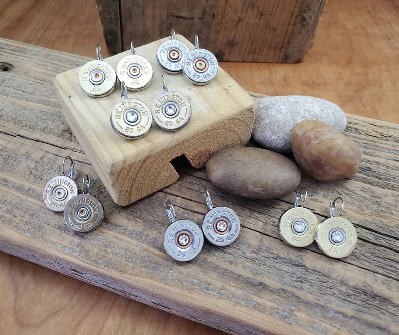May include: A set of silver and gold tone earrings featuring a round, silver-coloured shotgun shell with a clear rhinestone in the centre. The earrings are displayed on a wooden surface with small rocks.