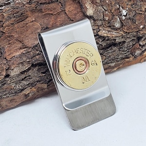 May include: A silver-colored money clip with a Winchester 12 GA shell casing. The brass casing is centered on the clip and features the text "WINCHESTER" and "GA". The clip is resting on a piece of brown wood.