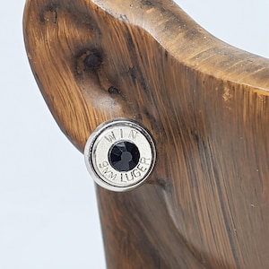May include: A pair of silver bullet earrings with a black gemstone. The earrings are engraved with "WIN 9MM LUGER".