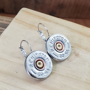 May include: A pair of silver hoop earrings with a silver and copper shotgun shell charm. The charm has the text "REMINTON 20 GA" on it.