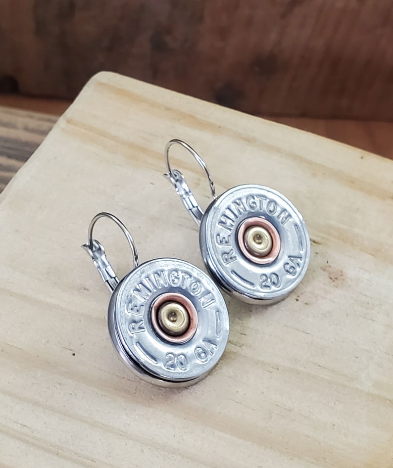 May include: A pair of silver hoop earrings with a silver and copper shotgun shell charm. The charm has the text "REMINTON 20 GA" on it.