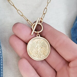 May include: Gold chain necklace with a toggle clasp and a round pendant featuring an embossed image of an angel with wings.