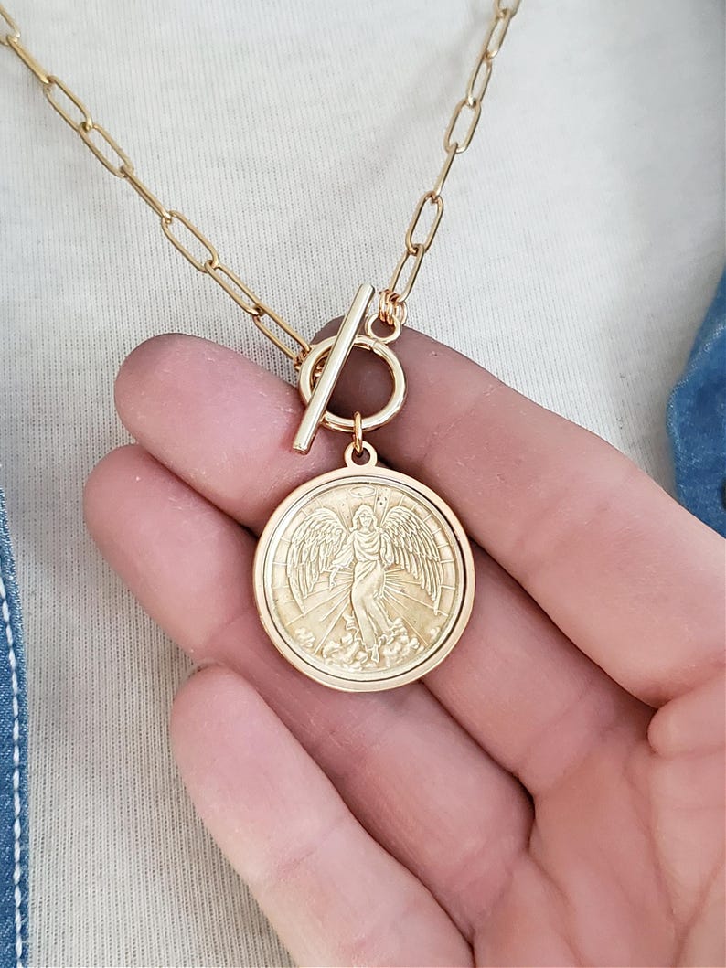 May include: Gold chain necklace with a toggle clasp and a round pendant featuring an embossed image of an angel with wings.