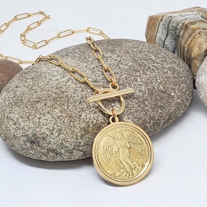 May include: Gold chain necklace with a toggle clasp and a large round pendant featuring an angel with wings. The pendant has a textured surface and a raised design.