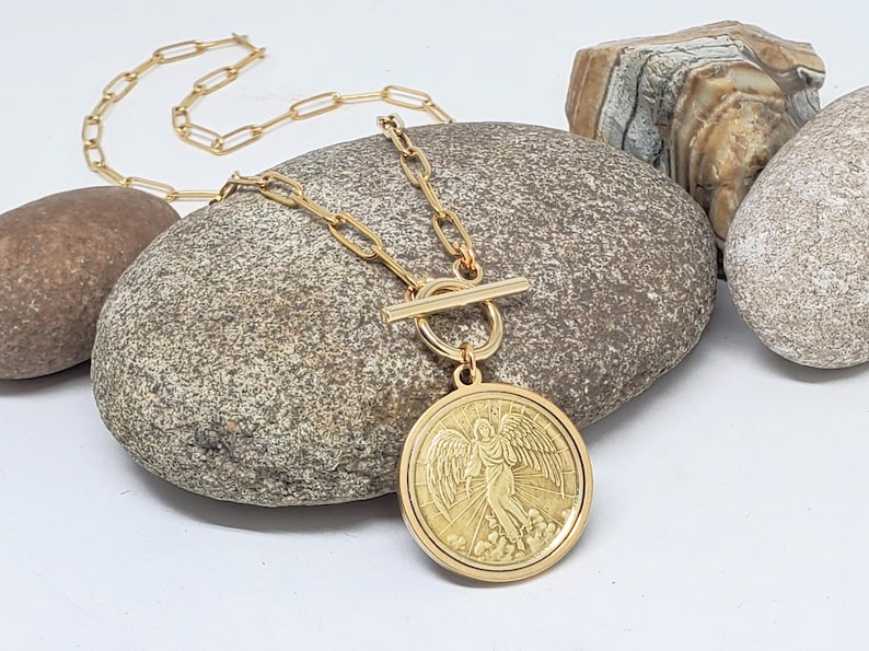 May include: Gold chain necklace with a toggle clasp and a large round pendant featuring an angel with wings. The pendant has a textured surface and a raised design.