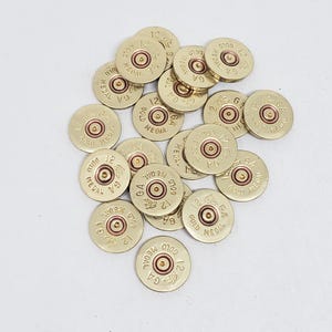 May include: A collection of brass-colored 12 GA Gold Medal shotgun shell casings. Each casing has the text "12 GA GOLD MEDAL" stamped on it. The casings are scattered across a white surface.