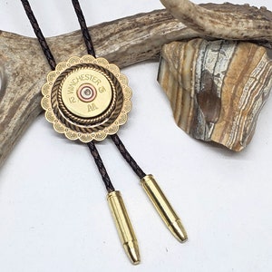Op de afbeelding: Een gouden bolo tie met een decoratieve messing behuizing rond een Winchester 12 GA-huls. De bolo tie heeft bruine leren koorden.