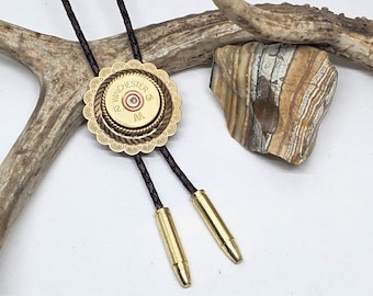 Puede incluir: Un bolo tie dorado con una carcasa de latón decorativa alrededor de un casquillo de escopeta Winchester 12 GA. El bolo tie tiene cordones de cuero marrón.