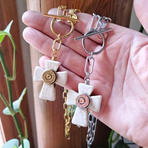 May include: Two necklaces featuring white stone crosses with brown veining, each centered with a metallic bullet casing. One necklace has a gold-tone chain and toggle clasp, while the other has a silver-tone chain and toggle clasp.