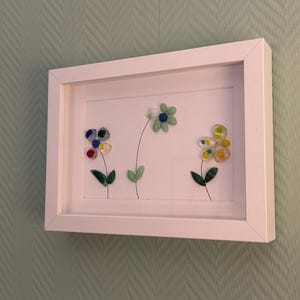 Könnte beinhalten: Ein weiß gerahmtes Kunstwerk mit drei bunten Glasblumen. Die Blumen sind in Blau-, Gelb- und Grüntönen gehalten und haben grüne Blätter und Stiele. Sie sind vor einem weißen Hintergrund platziert. Das Kunstwerk hängt an einer hellgrünen Wand.