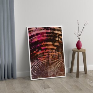 May include: Framed abstract art print with a white border. The artwork features a geometric design with concentric circles and layered shapes in shades of red, orange, and brown. A small, red vase with branches sits on a wooden stool.