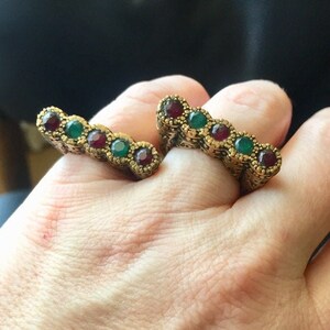 May include: Two gold rings with red and green gemstones. The rings are worn on the index and middle fingers of a hand.