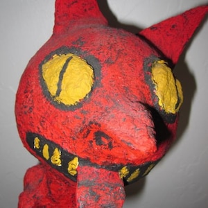 May include: A red papier-mâché figure with yellow eyes and a black painted mouth. The figure has a long, pointed nose and a tongue sticking out. The figure is painted with black and red paint and has a rough texture.
