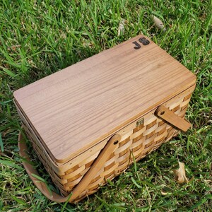 May include: A wooden picnic basket with a hinged lid and a woven wicker base. The lid has a natural wood finish and the initials "JS" are engraved on the top. The basket has two wooden handles.