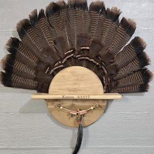 May include: A mounted turkey fan display. The fan is composed of brown and black feathers, mounted on a wooden base. The base has a shelf with the inscription "Kansas 4/15/17". A decorative element with beads and a black tassel hangs below.