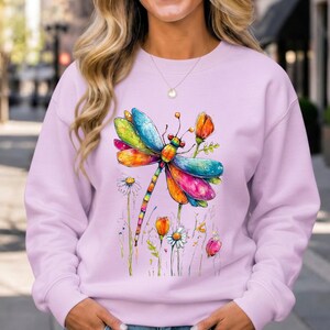May include: A light purple long-sleeve sweatshirt featuring a colorful watercolor-style graphic of a dragonfly surrounded by various flowers. The dragonfly has vibrant wings in shades of blue, yellow, orange, and pink, with a long, segmented body.