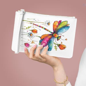 May include: A white rectangular wristlet pouch featuring a colorful watercolor illustration of a dragonfly with vibrant wings in shades of red, yellow, blue, and green, surrounded by small painted flowers and thin stems on a plain white background.